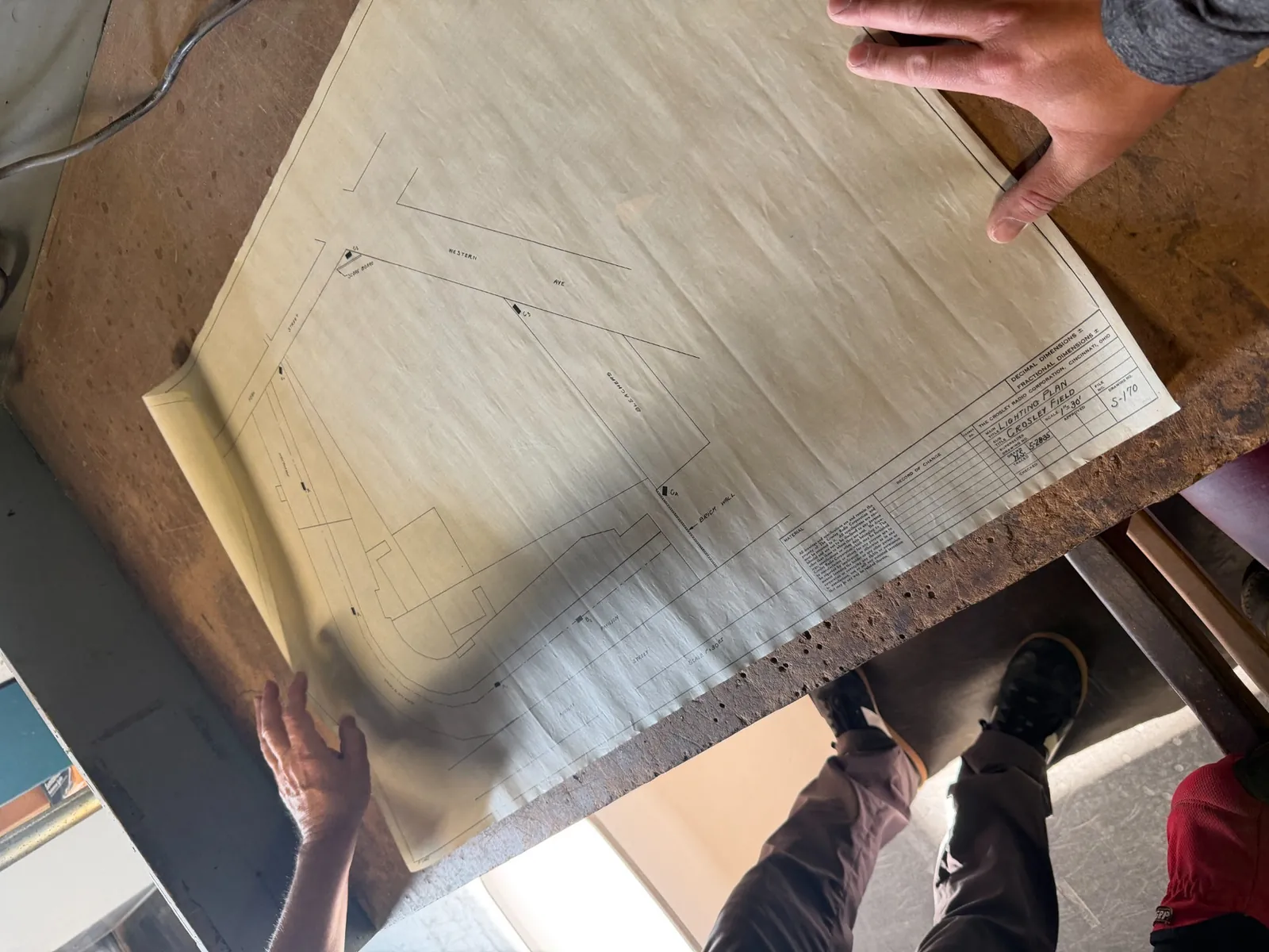 Original-period engineering drawing labeled Lighting Plan Crosley Field spread across a worktable, with hands holding the corners